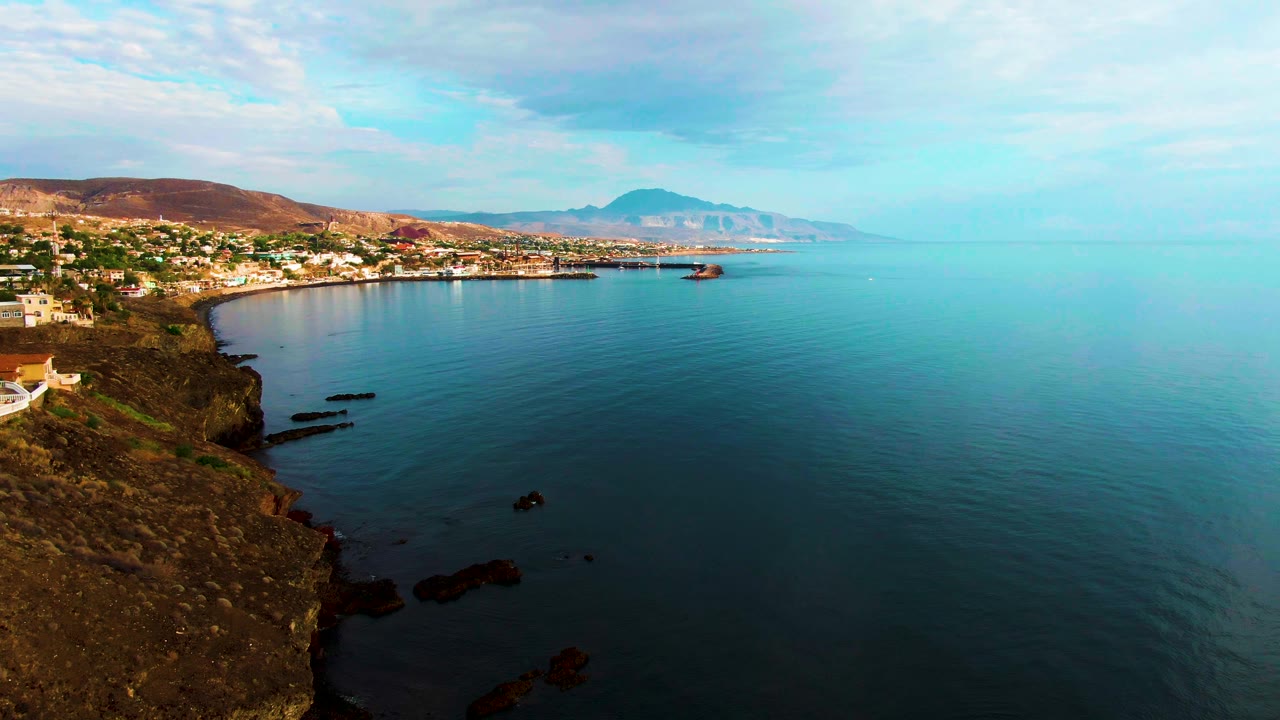 Rocky headland beside a coastal town with calm blue water and distant mountains.
