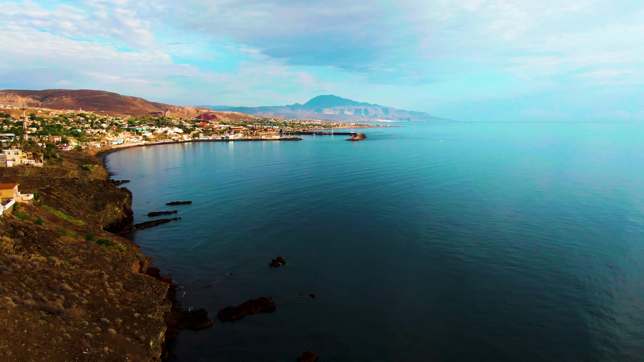 Santa Rosalia coastal settlement lines the left side below brown hills, with rocky shoreline below, turquoise-blue sea across the frame, soft cloud cover, and no visible people.