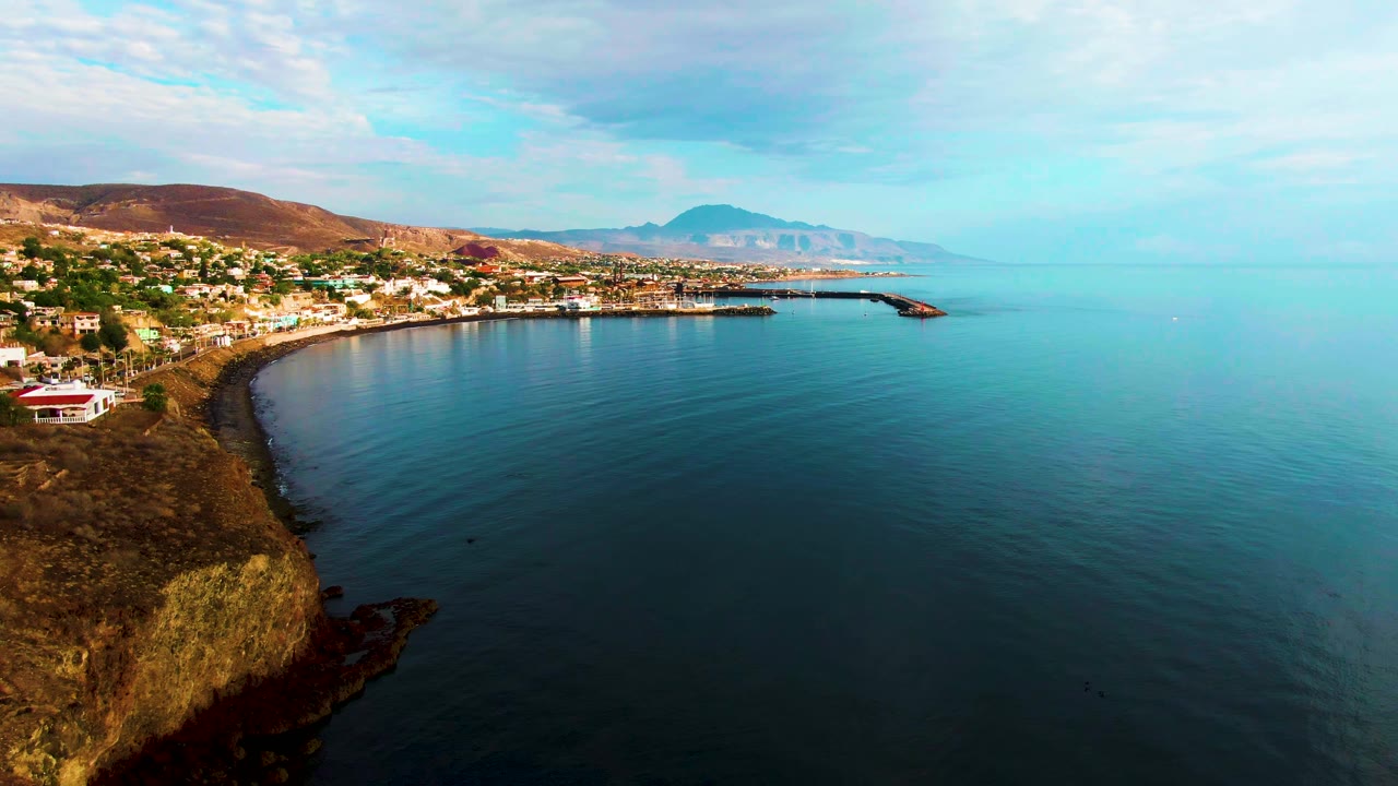 Aerial view over Santa Rosalia, Baja California Sur, with a rocky cliff at lower left, a curved blue bay, shoreline buildings, distant hills, pale sky, and no visible people.