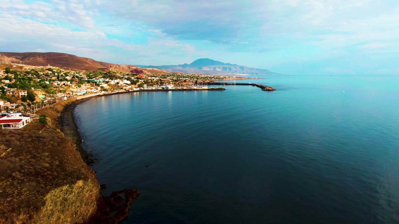 Wide aerial frame over Santa Rosalia's curved shoreline, with open turquoise water on the right, the town stretching along the left coast, brown hills behind it, and no visible people.