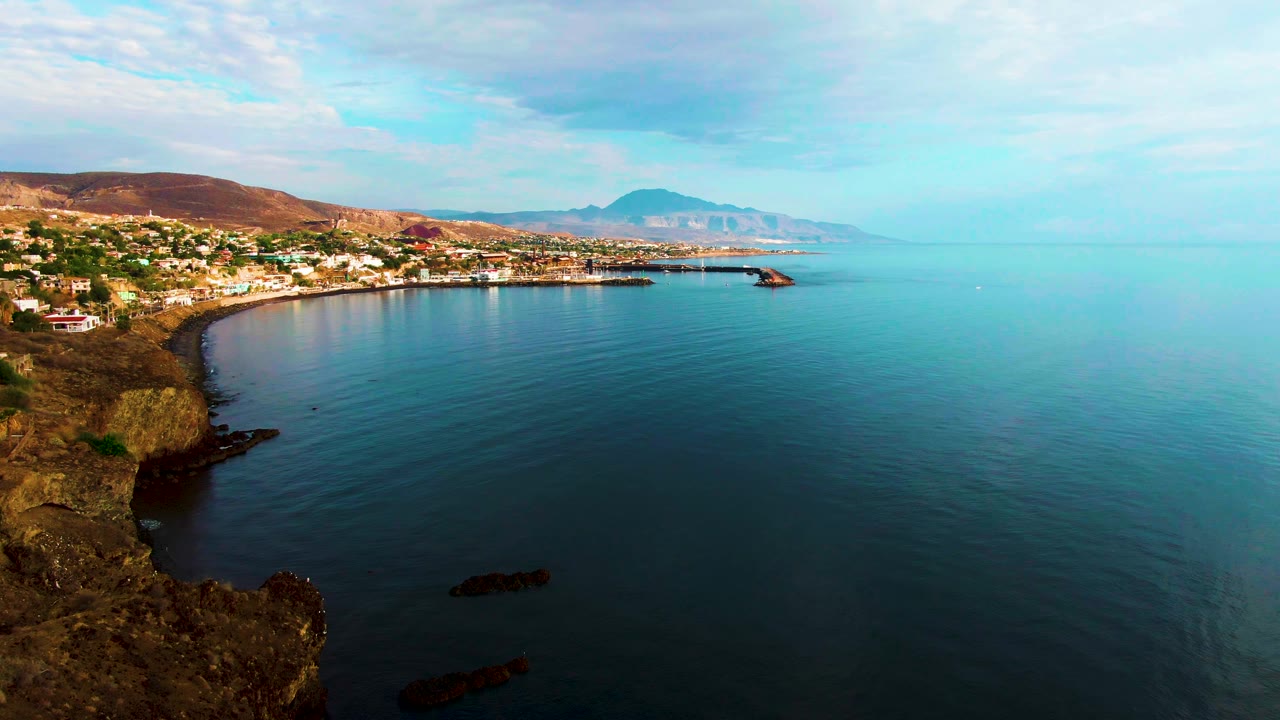 Santa Rosalia, Baja California Sur appears from above with rocky cliffs in the foreground, shoreline buildings to the left, calm blue-green ocean to the right, distant hills, and no visible people.