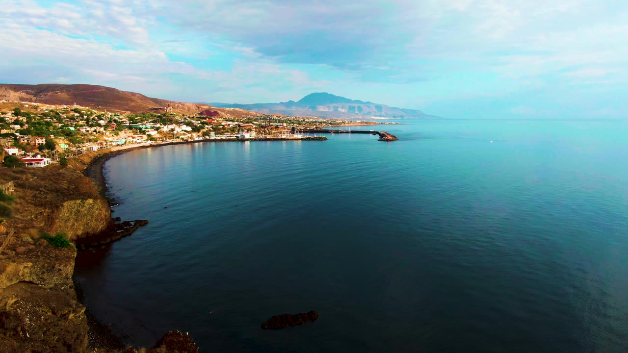Santa Rosalia's coastal strip sits along the left side of the frame, with brown cliffs below, calm water spreading across the right, a pale sky above, and no visible people.
