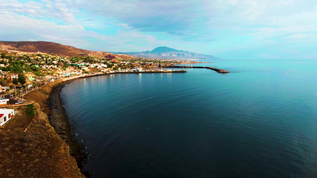 Opening aerial frame over Santa Rosalia, Baja California Sur, Mexico, with the cliff edge at lower left, a curved rocky shoreline, compact town buildings, harbor breakwaters, blue-green sea, distant hills, and no visible people.