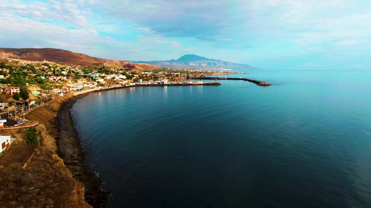 Aerial view at 0:01 over Santa Rosalia with more open sea on the right, shoreline buildings across the left, a harbor wall in the distance, dry hills behind town, and no visible people.