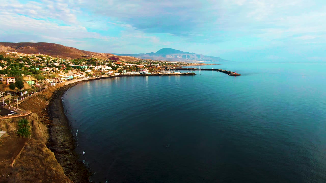Aerial frame at 0:02 showing Santa Rosalia's curved bay from above, with brown cliffside terrain along the near shore, town buildings on the left, a narrow harbor structure ahead, calm sea, and no visible people.