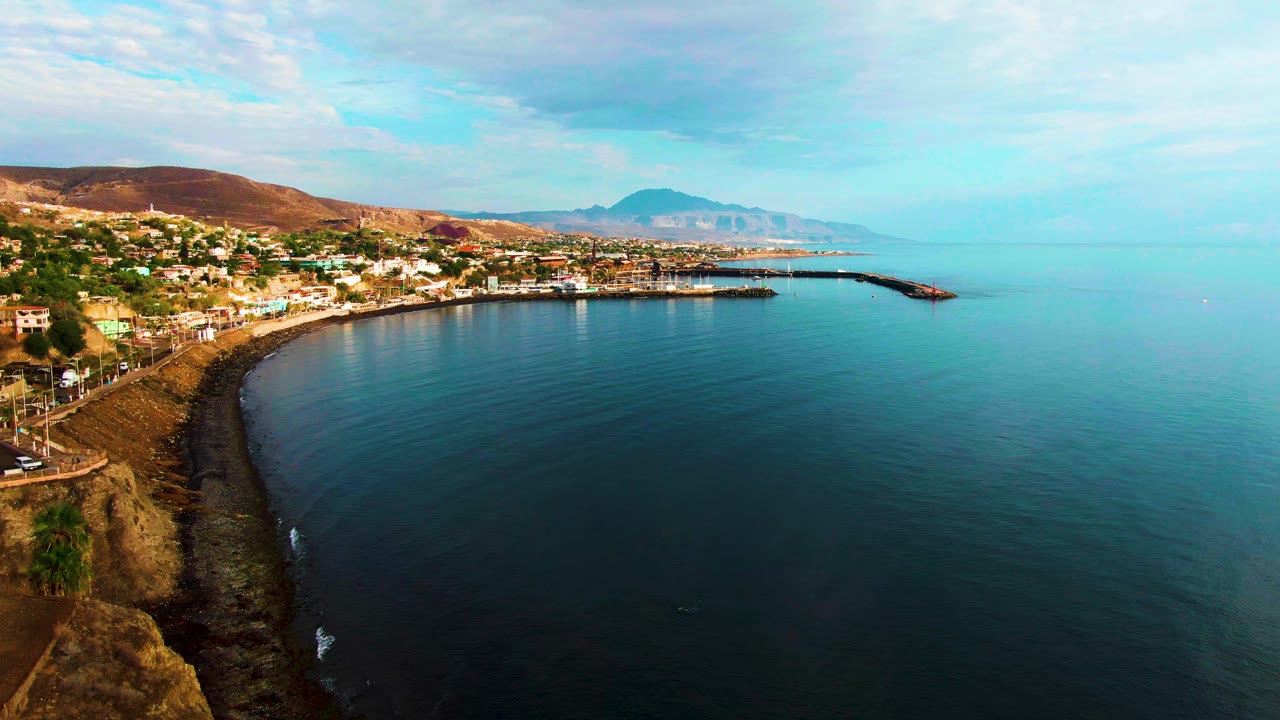 Aerial coastline view with rocky cliff foreground, town buildings, harbor, and open sea.