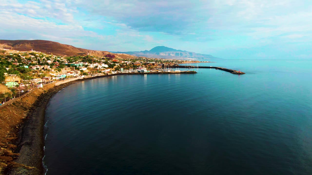 Opening aerial frame over Santa Rosalia, Baja California Sur, with a curved rocky shore, dark calm water, town buildings, hills, distant mountains, scattered clouds, and no visible people.
