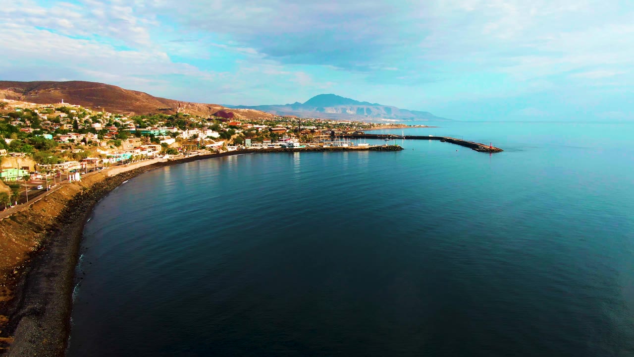 Second aerial frame in Santa Rosalia showing the shoreline slightly farther along the curve, with buildings on the left, harbor breakwaters near center, open ocean to the right, and no visible people.