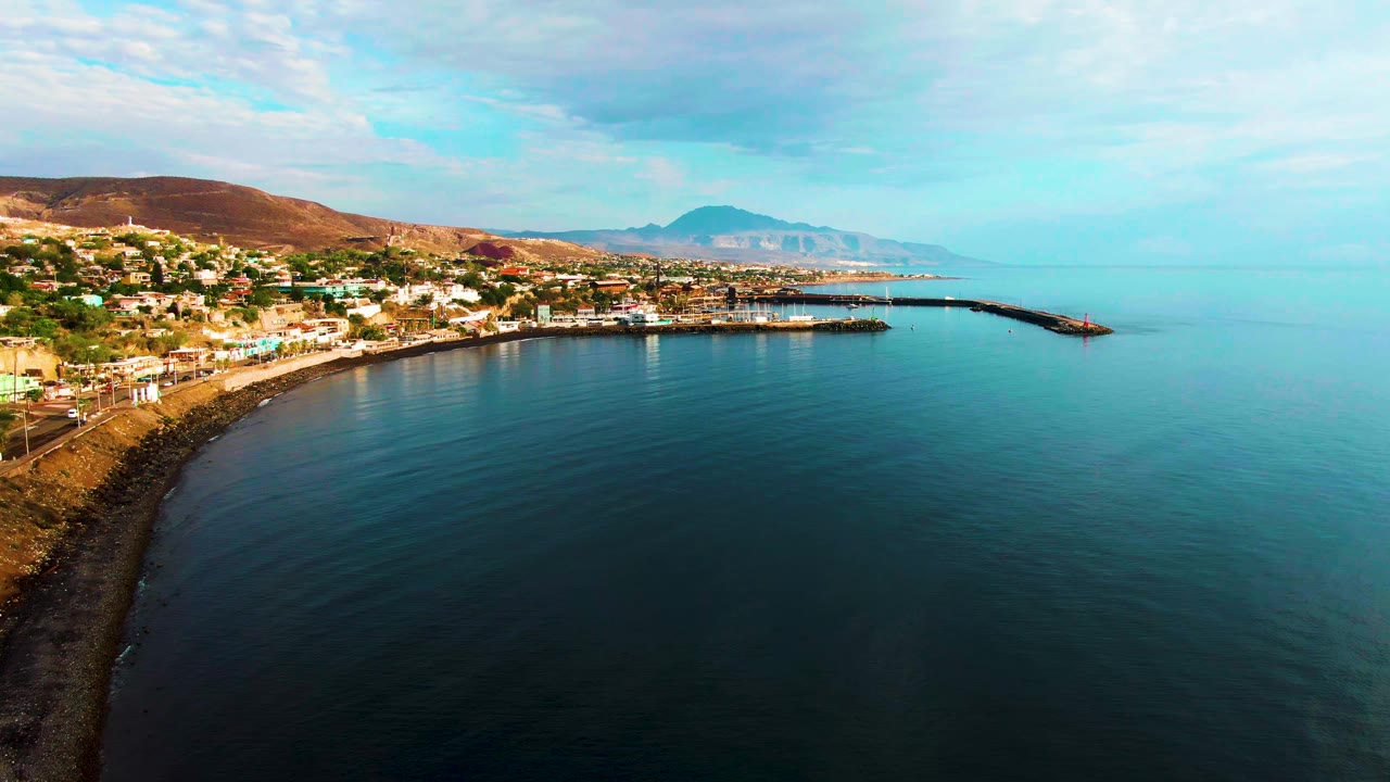 Third frame holds a high drone view of Santa Rosalia's coast, with the town along the left shoreline, a narrow harbor projection, smooth blue-green sea, hazy mountains, and no visible people.