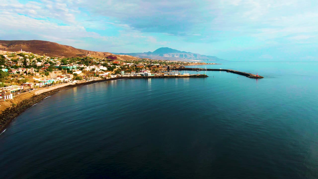 Opening aerial frame over Santa Rosalia, Baja California Sur, with dark calm ocean in the foreground, a curved shoreline town at left, a harbor pier reaching into the sea, distant mountains, and no visible people.