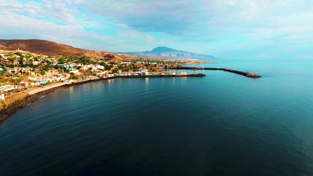 Wide drone view of shoreline buildings, harbor pier, calm sea, hills, and distant mountains.