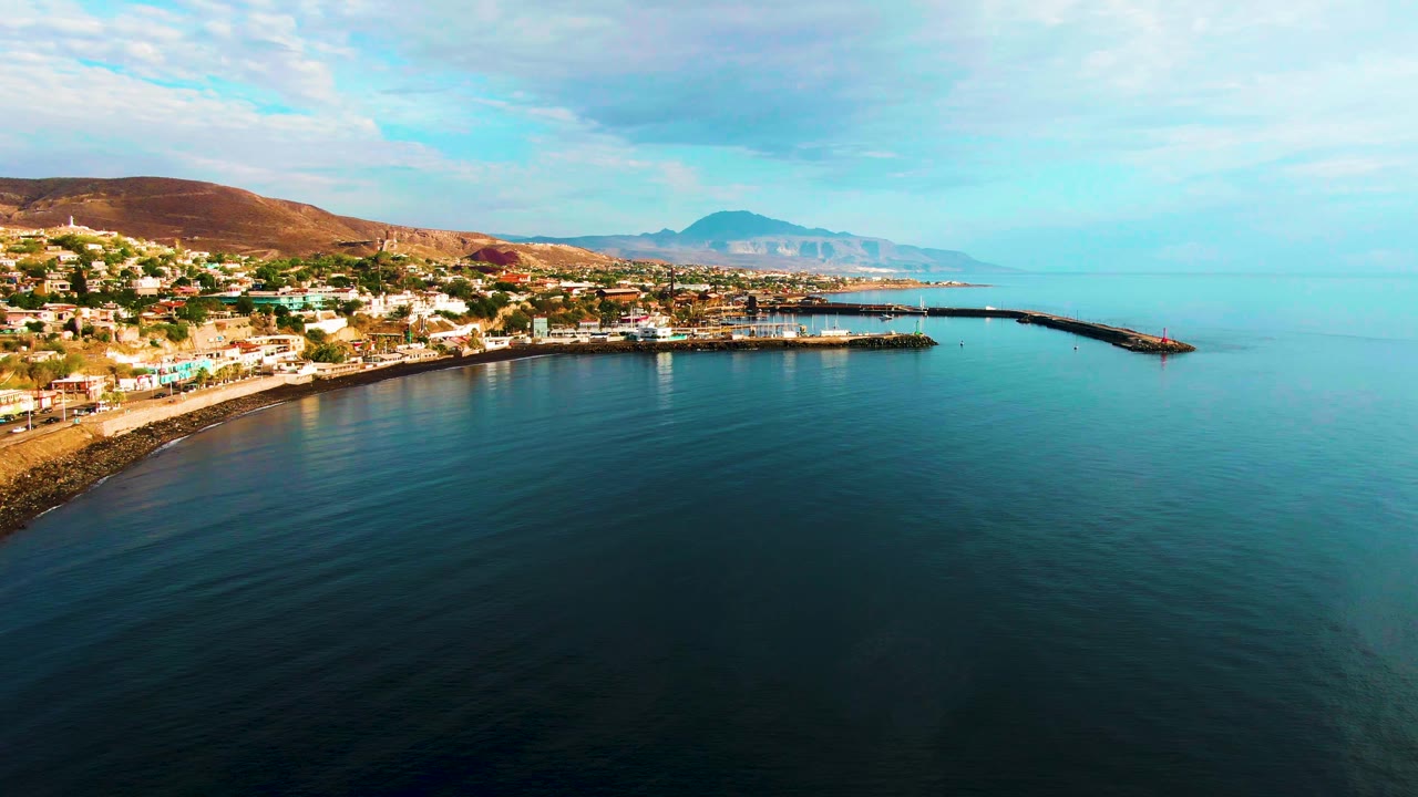 Aerial frame of Santa Rosalia showing the town along the left shoreline, the pier extending toward the right, deep blue water in the foreground, low hills behind the buildings, and no visible people.