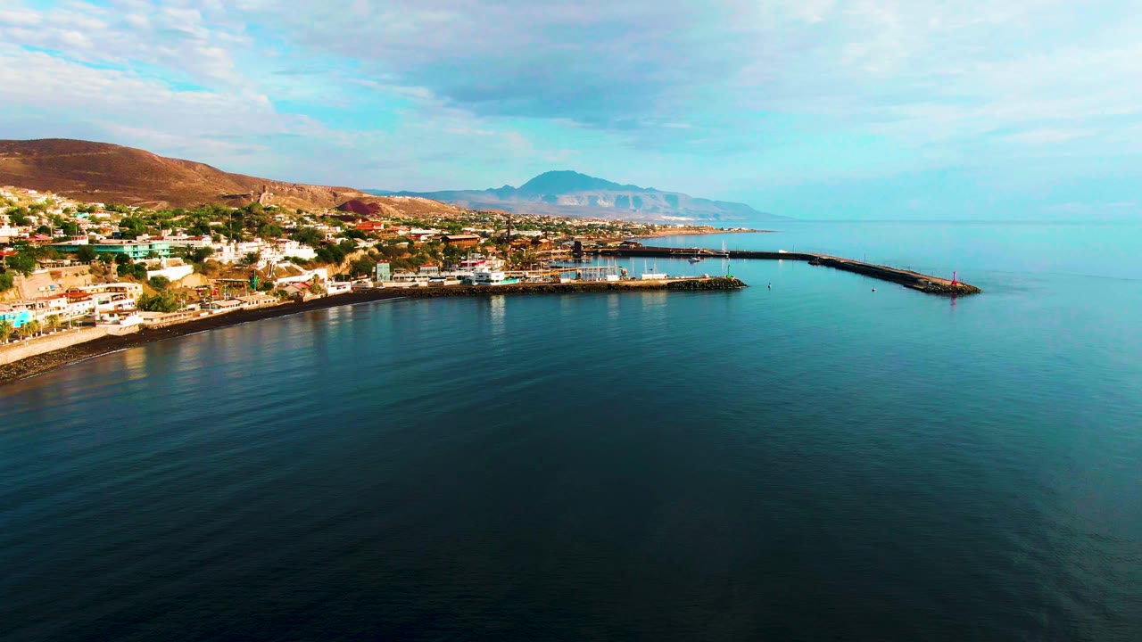 Opening aerial frame over Santa Rosalia, Baja California Sur, with dark rippled ocean in front, the town and harbor at left, a long breakwater to the right, distant mountains, and no visible people.
