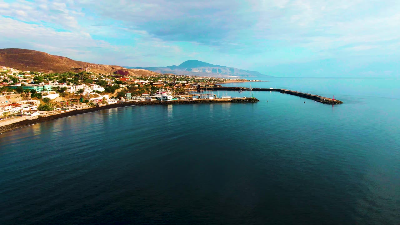 Aerial view one second into the Santa Rosalia clip, with the breakwater extending across the mid-right water, hillside buildings along the left shore, soft clouds overhead, and no visible people.