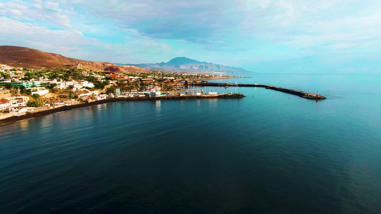 Wide aerial frame over Santa Rosalia showing broad blue-green water in the foreground, the town compressed along the left coast, the breakwater reaching toward the right edge, mountains in haze, and no visible people.