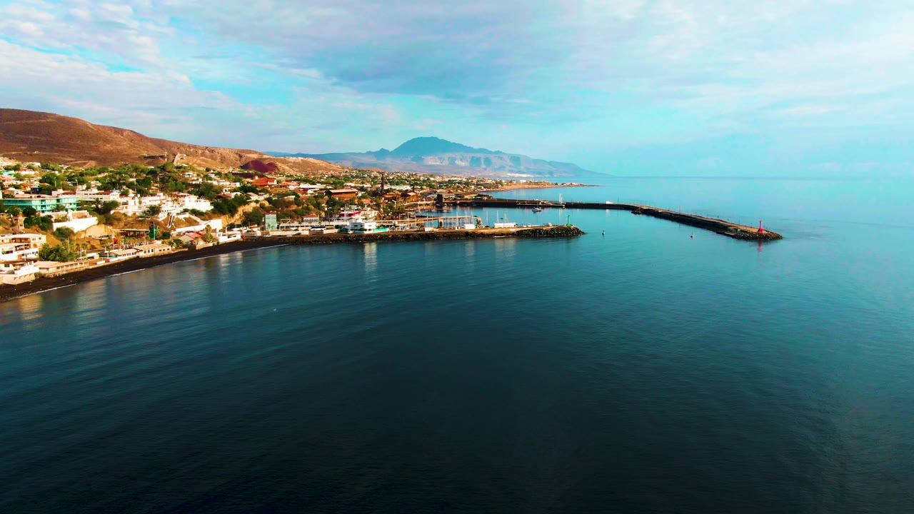 Aerial coastal town and harbor beside calm ocean under scattered clouds.