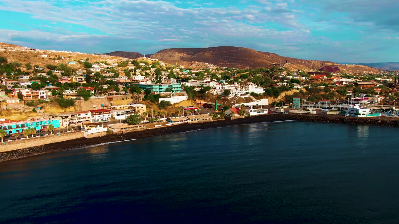 Opening frame of Santa Rosalia, Baja California Sur, showing calm dark blue water below a waterfront strip, hillside buildings, brown mountains, partly cloudy sky, and no visible people.