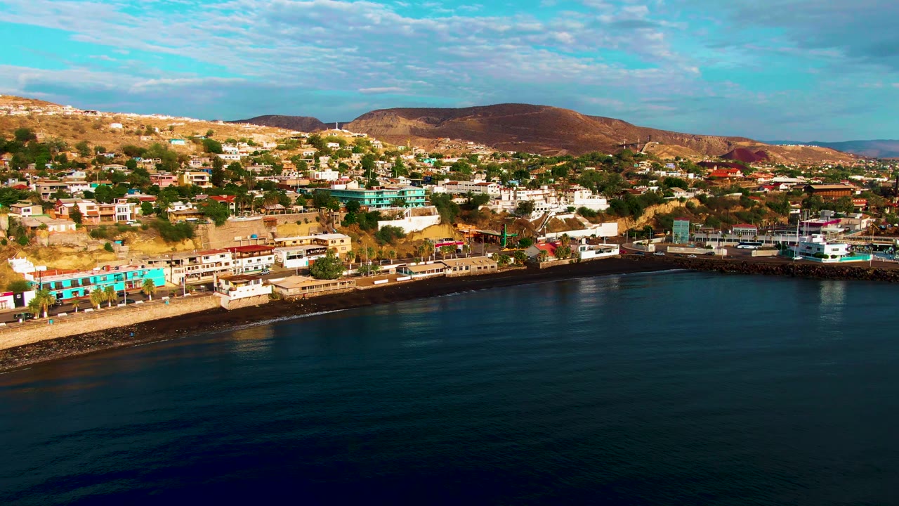 Second frame of Santa Rosalia with the shoreline slightly farther across the view, showing blue-green waterfront buildings, hillside housing, dry hills, calm sea, and no visible people.