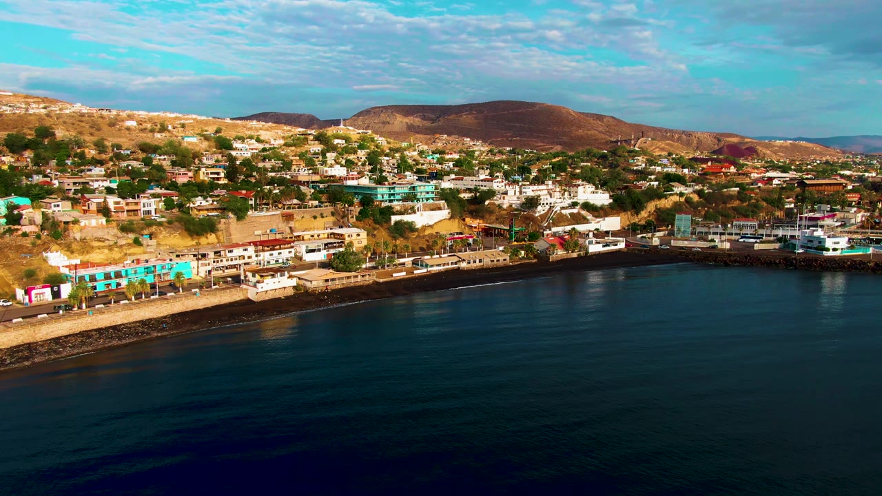 Third frame of Santa Rosalia showing the waterfront curve, dark calm water in the foreground, dense hillside buildings, arid mountain ridges, scattered clouds, and no visible people.