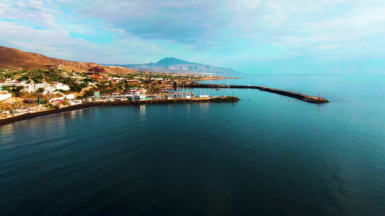 Opening frame of Santa Rosalia harbor in Baja California Sur, with dark blue water in the foreground, a curved rocky pier, shoreline buildings, brown hills, distant mountains, and no visible people.