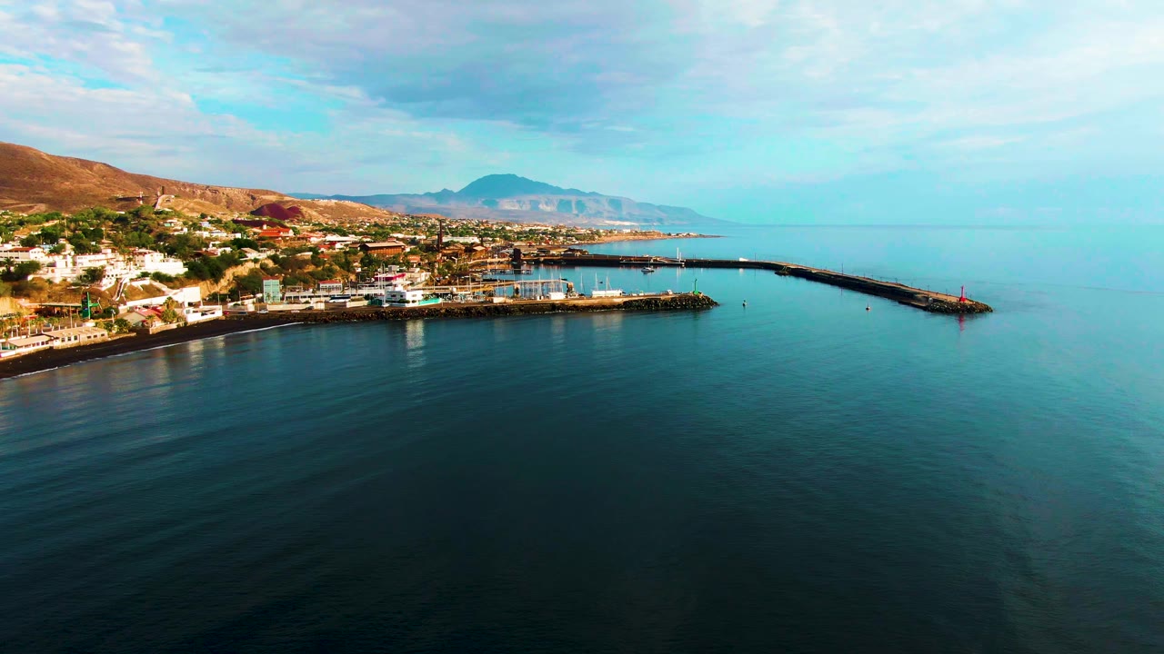 Wide coastal harbor view with pier, small waterfront town, calm water, and mountains.