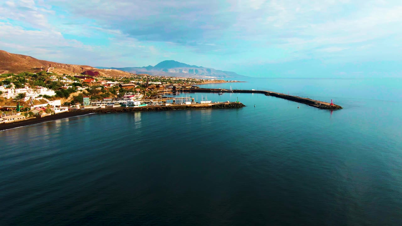 Aerial frame of Santa Rosalia's waterfront with open sea filling the foreground and right side, the long pier cutting across the harbor, town buildings at left, distant mountains, and no visible people.