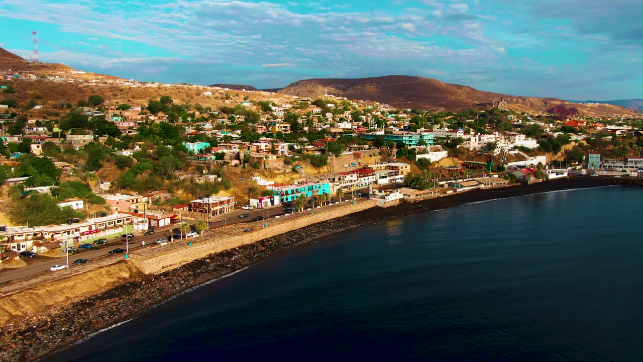 Aerial frame of Santa Rosalia, Baja California Sur with a waterfront road beside dark calm water, low buildings below dry hills, and no visible people.