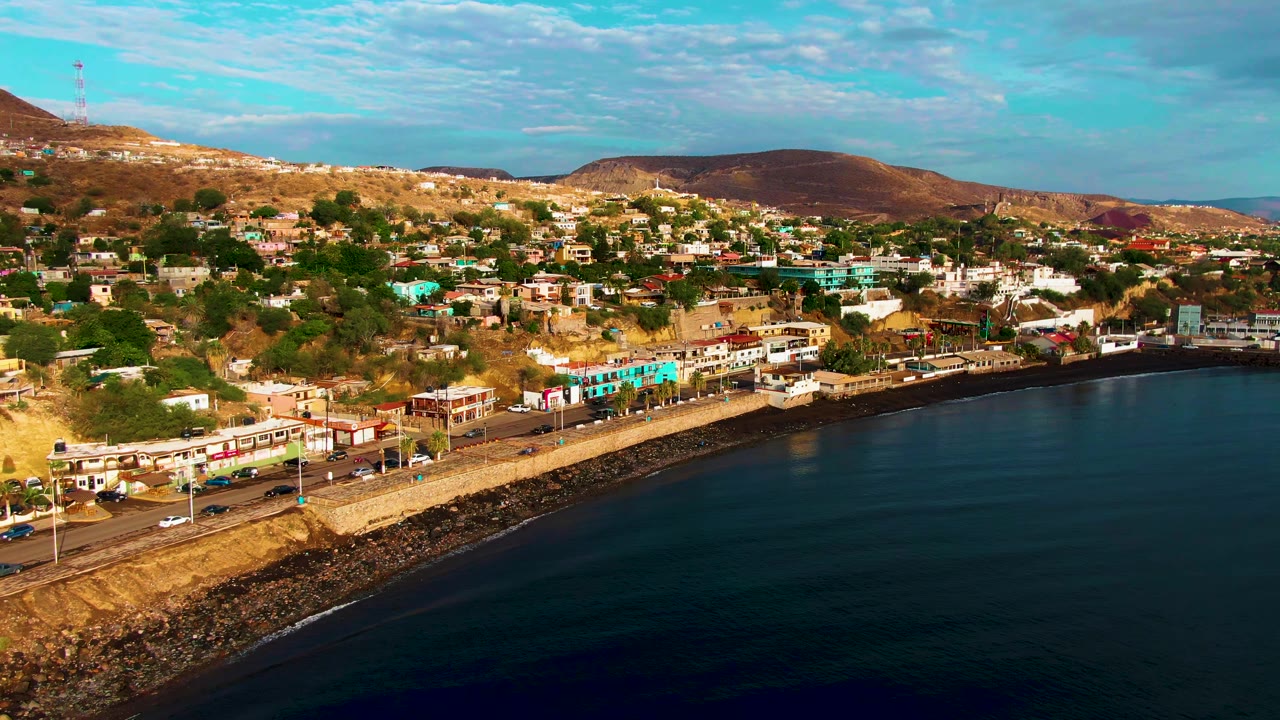 Aerial view over Santa Rosalia showing cars on the shore road, clustered waterfront buildings, dark ocean water, brown hills, and no visible people.