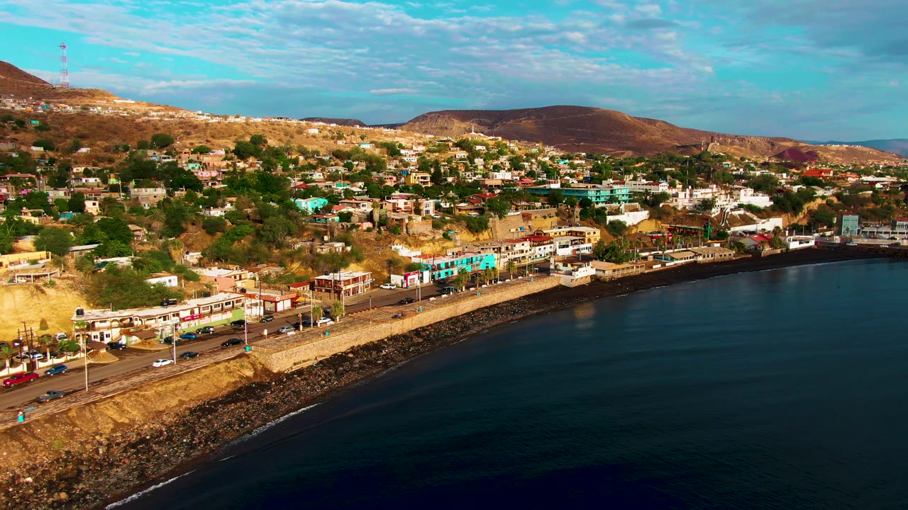 Aerial shoreline view with vehicles, buildings, ocean, and hills.