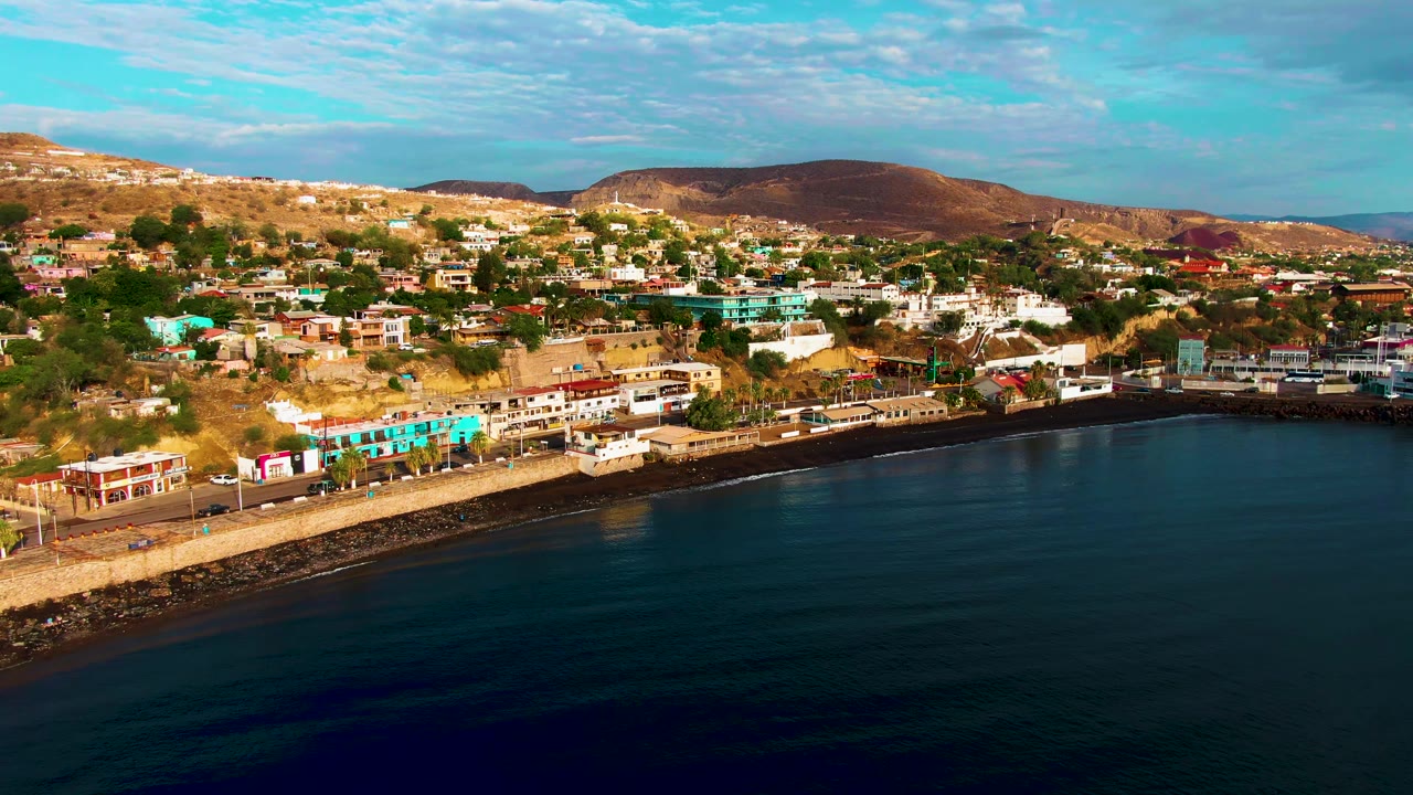 Aerial view over Santa Rosalia, Baja California Sur, with dark blue water in the foreground, a seawall road below hillside buildings, brown hills beyond, and no visible people.