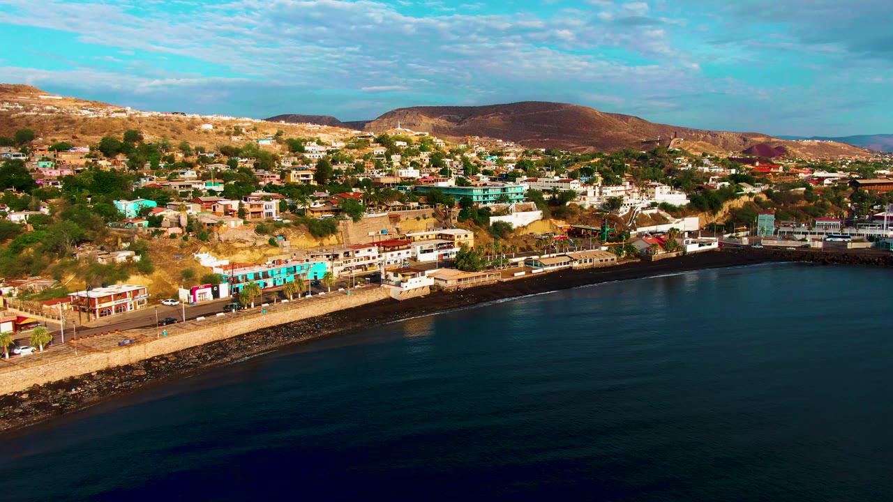 Santa Rosalia shoreline road curves beside dark water, with small vehicles on the highway, colorful hillside buildings above it, distant brown hills, and no visible people.
