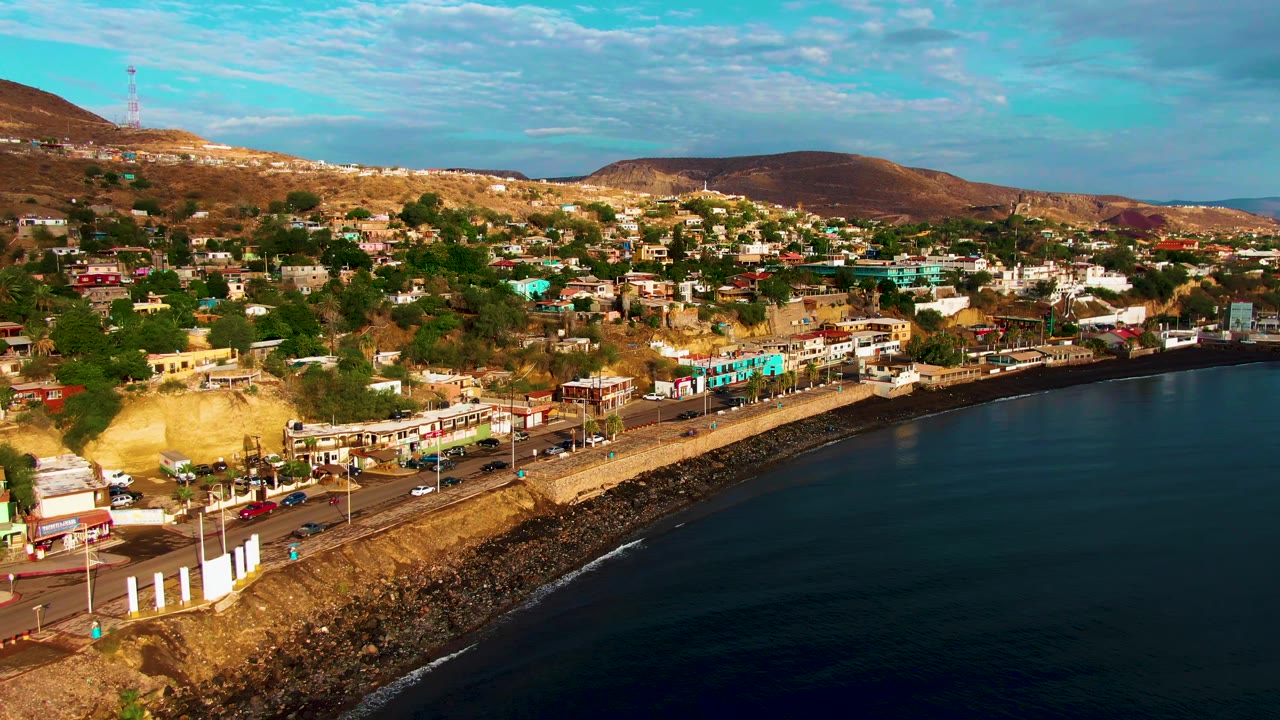 Aerial view of a coastal road, ocean, town buildings, and dry hills.