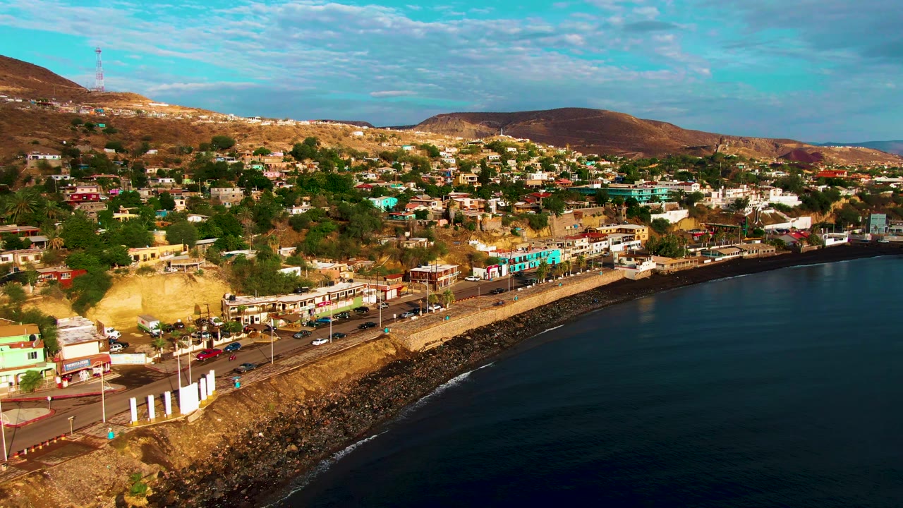 Aerial view in Santa Rosalia showing the shoreline road closer to the lower edge, turquoise and white buildings near the waterfront, scattered homes on the slope, and no visible people.