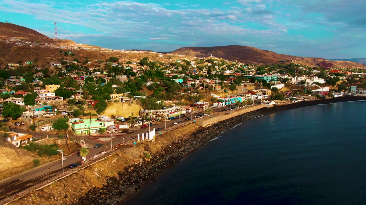 One second into the clip, Santa Rosalia's shoreline road runs along the lower frame with vehicles, hillside homes and trees above it, dark bay water at right, and no visible people.