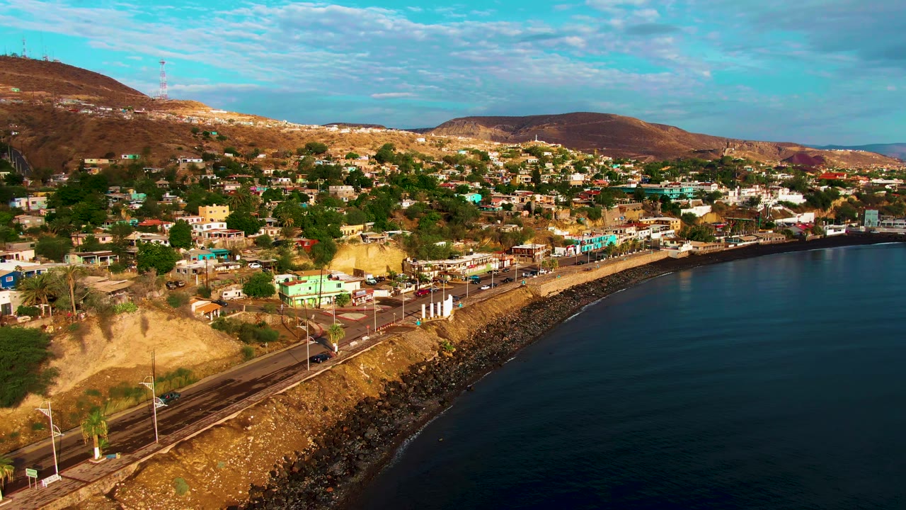 Opening aerial view in Santa Rosalia, Baja California Sur, with a rocky shore road, hillside buildings, brown hills, blue water, and no visible people.