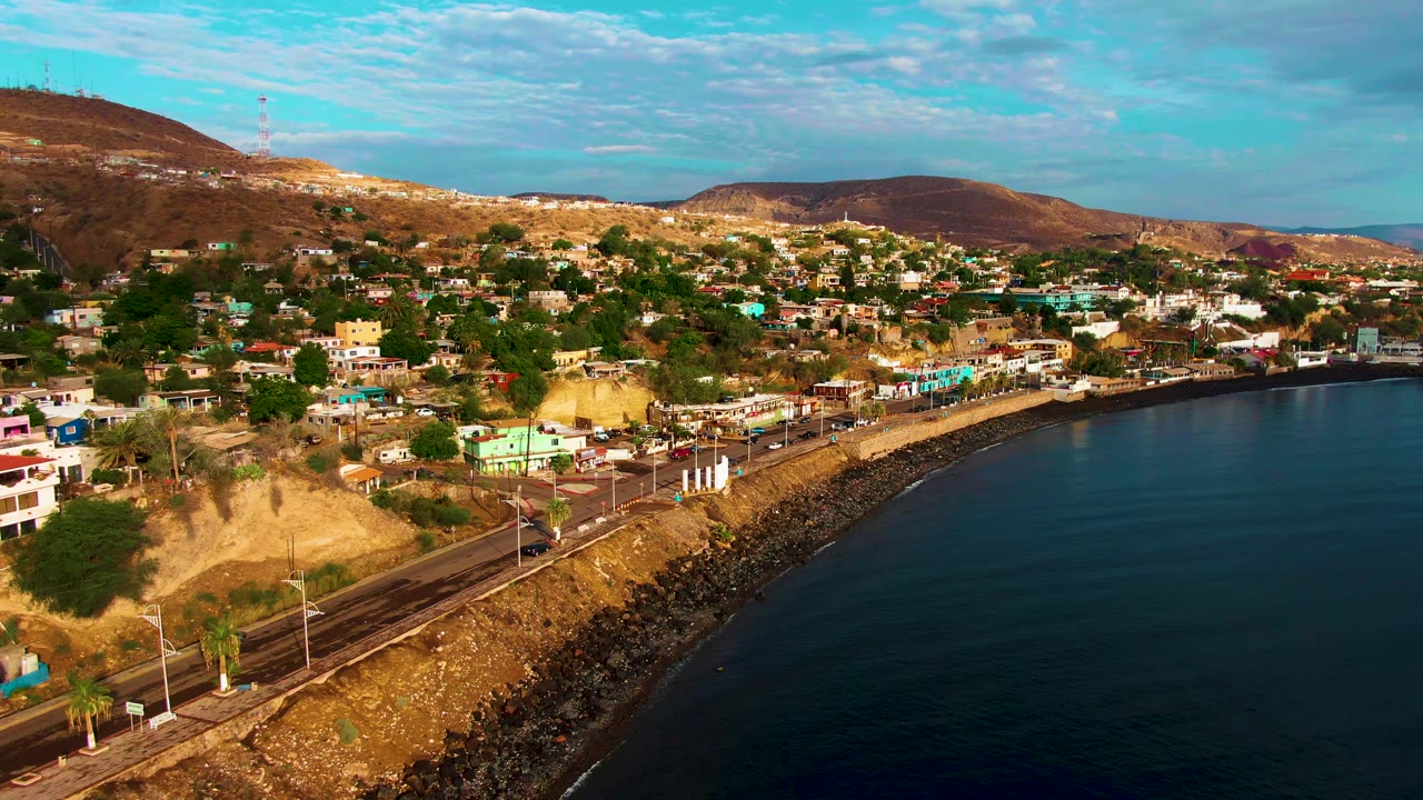 Aerial frame in Santa Rosalia showing the shoreline road slightly farther along the curve, with vehicles, hillside homes, trees, brown hills, and no visible people.
