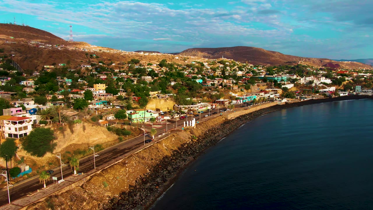 Wide aerial view of Santa Rosalia with the curved coastal road, dark water, hillside buildings, roadside poles, dry hills, and no visible people.