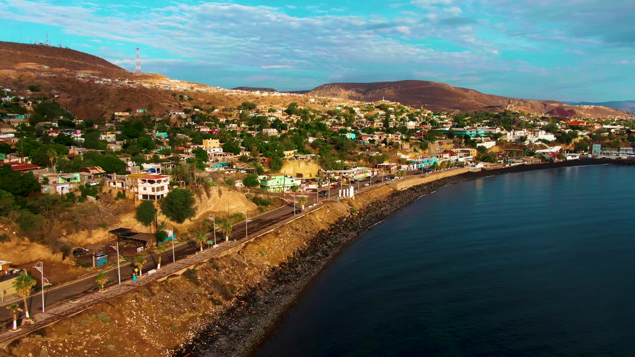 First aerial view of Santa Rosalia, Baja California Sur, with a curving coastal road, hillside homes, dry brown slopes, calm blue water, and no visible people.