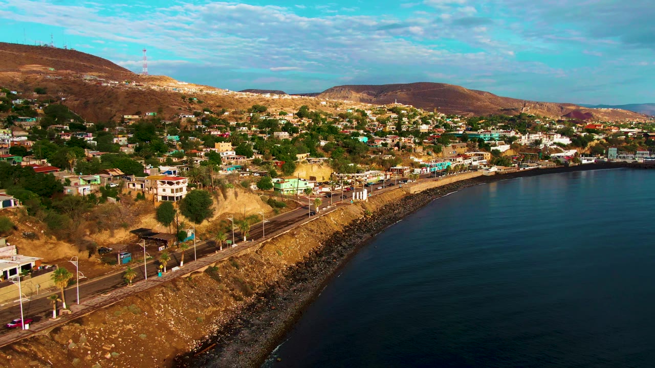 Aerial frame of Santa Rosalia showing vehicles along the shoreline road, clustered homes on the slope, rocky coast protection, blue water, and no visible people.