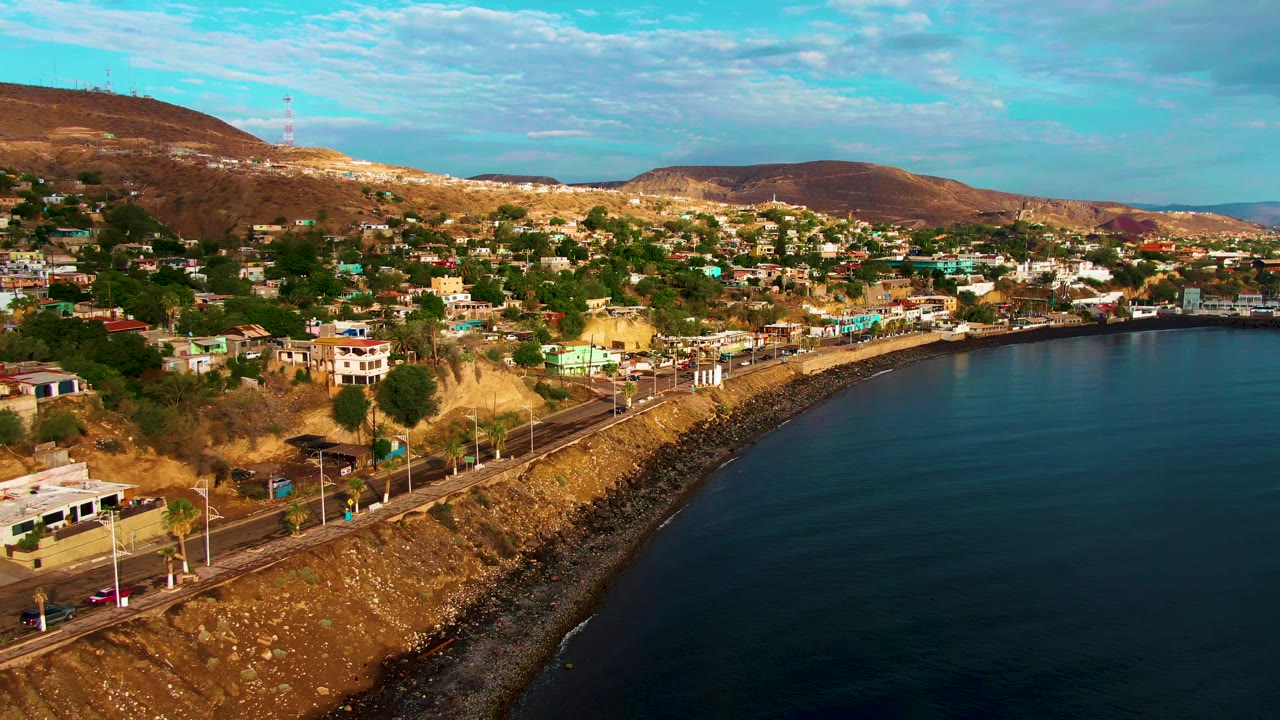 Santa Rosalia coastal road curves around the bay with cars and a bus visible, hillside residences above, brown ridges in back, deep blue ocean at right, and no visible people.