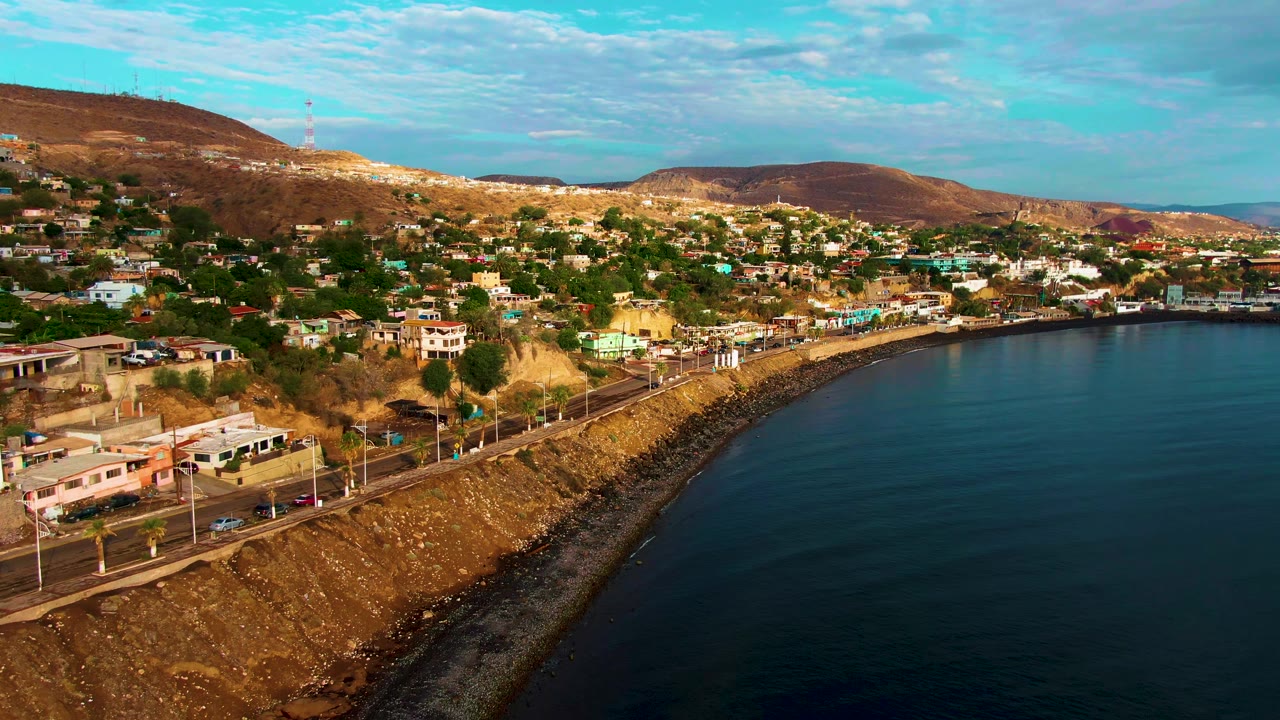 Aerial view of Santa Rosalia, Baja California Sur with a curving coastal road, hillside homes, dark ocean water at right, dry hills in the background, and no visible people.