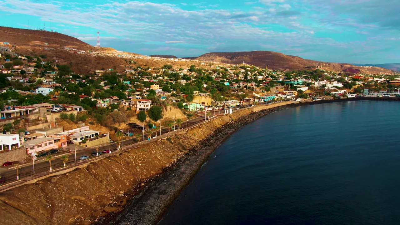 Santa Rosalia hillside neighborhood and waterfront road seen from above, with the bay on the right, scattered homes on dry slopes, and no visible people.