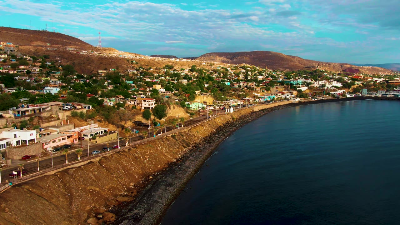 Aerial frame over Santa Rosalia with a road and streetlights along the shore, compact homes across the hillside, broad ocean water at right, and no visible people.