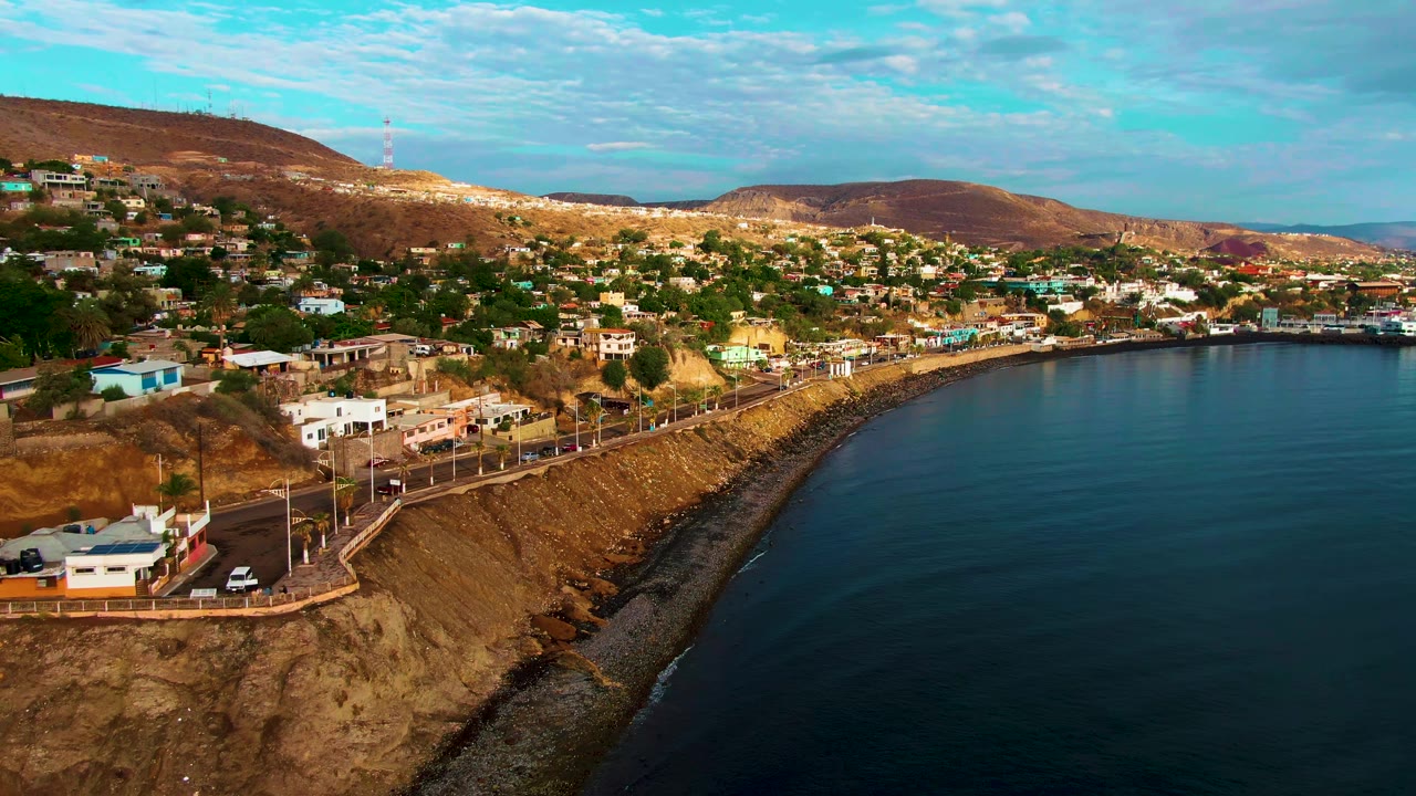 Aerial view in Santa Rosalia, Baja California Sur, with a coastal road on a cliff, hillside homes, calm dark blue ocean water, brown hills, and no visible people.
