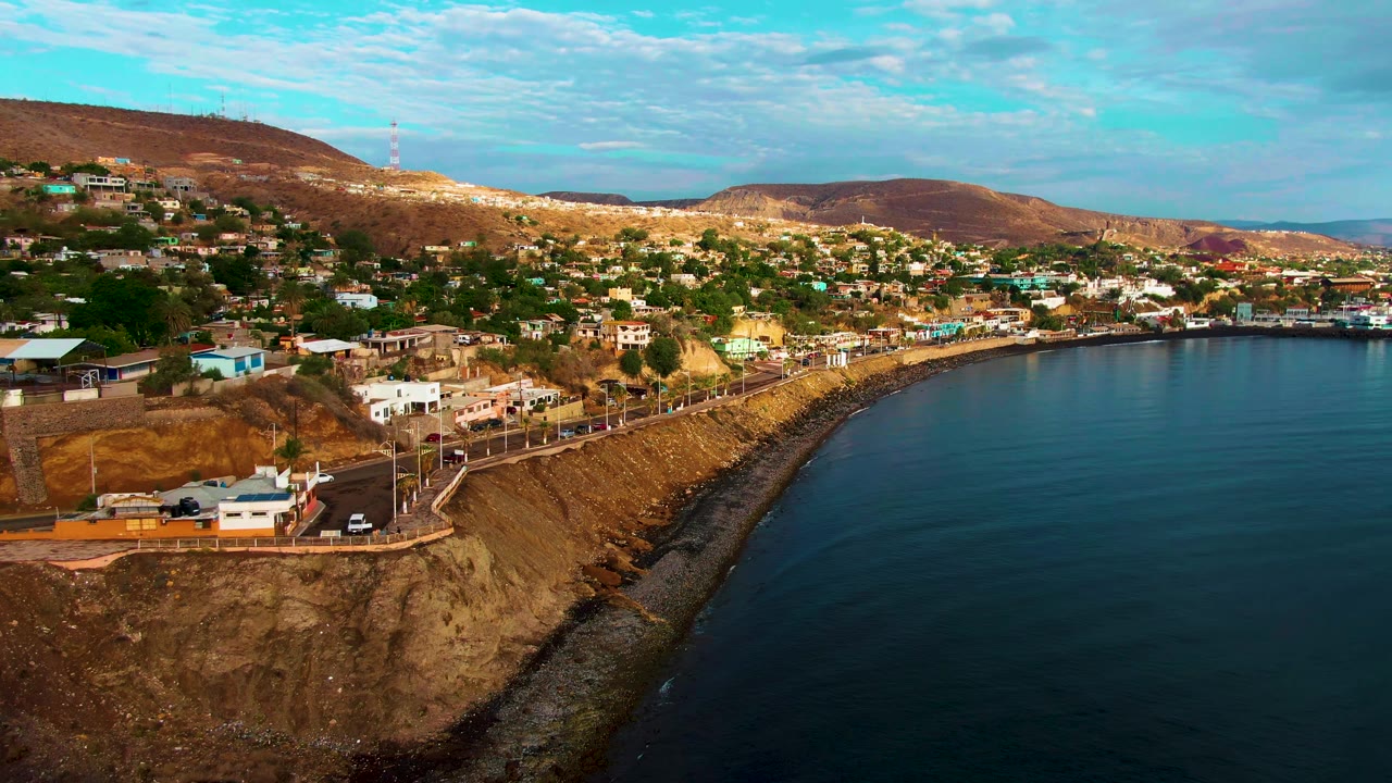 Aerial view over Santa Rosalia with the cliff road lower left, houses spread across the hillside, calm ocean filling the right side, distant hills, and no visible people.