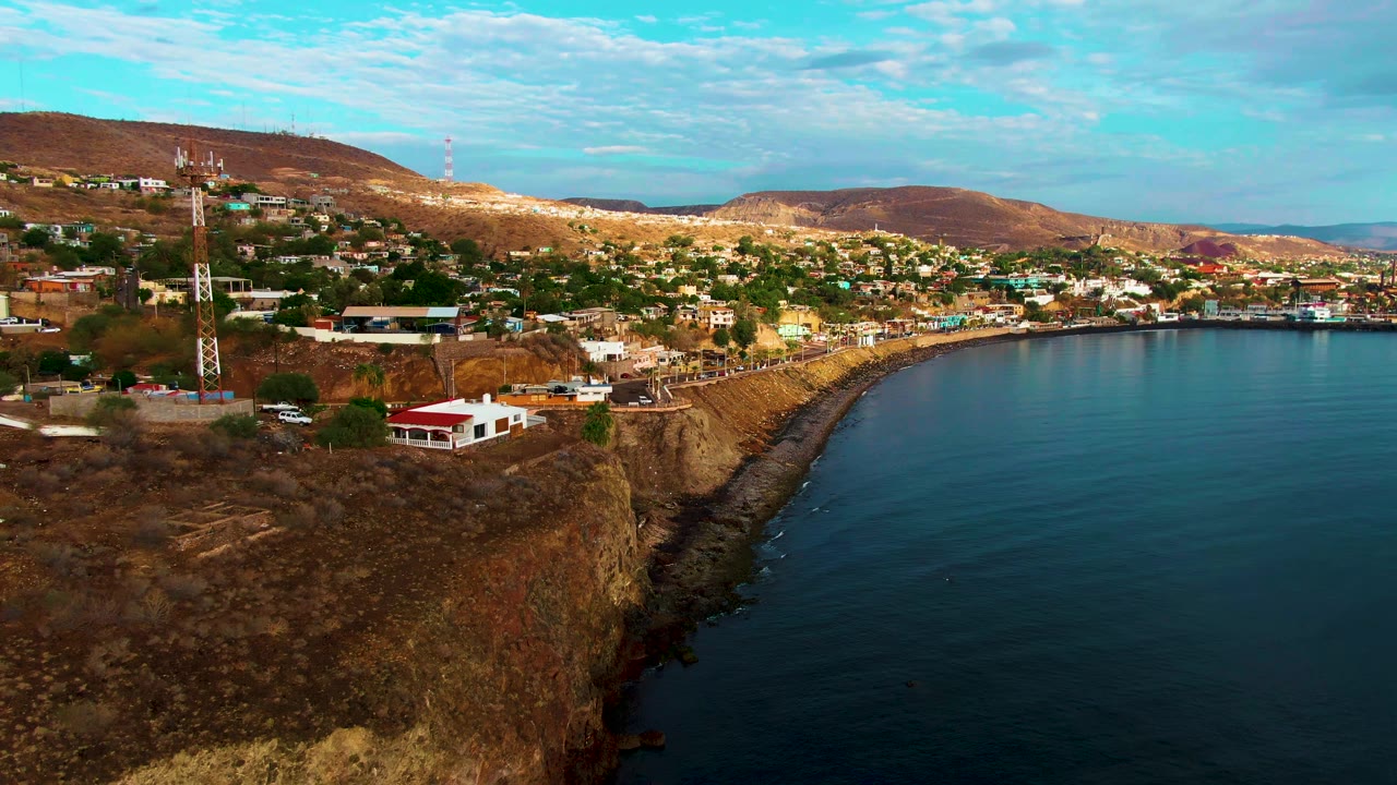 Opening aerial frame over Santa Rosalia, Baja California Sur, with a rocky cliff in the foreground, hillside houses and towers beyond it, blue ocean on the right, and no visible people.