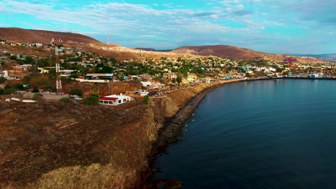 Coastal cliff with town buildings, towers, ocean water, and hills under clouds.