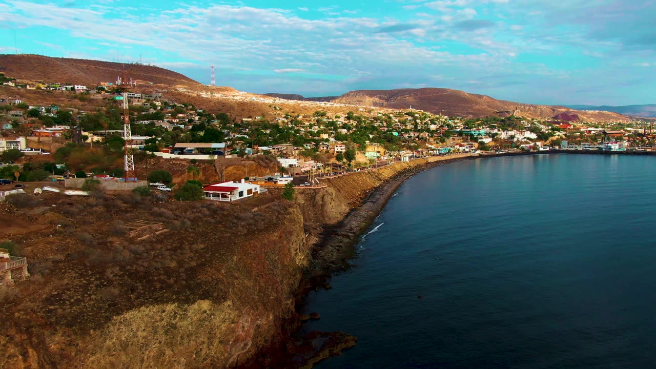 Santa Rosalia aerial view with the cliff dropping into dark blue water, town buildings spread across the hillside, brown mountains behind them, and no visible people.