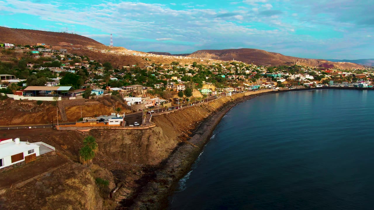 Aerial frame over Santa Rosalia, Baja California Sur, with hillside homes, a road along the cliff, blue water on the right, dry hills in back, and no visible people.
