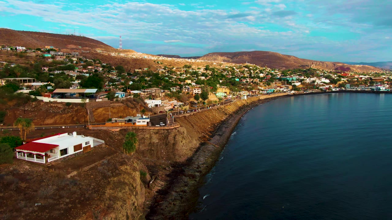 Aerial frame in Santa Rosalia with the red-roofed white building more centered on the cliff, small roadside vehicles nearby, calm water to the right, and no visible people.